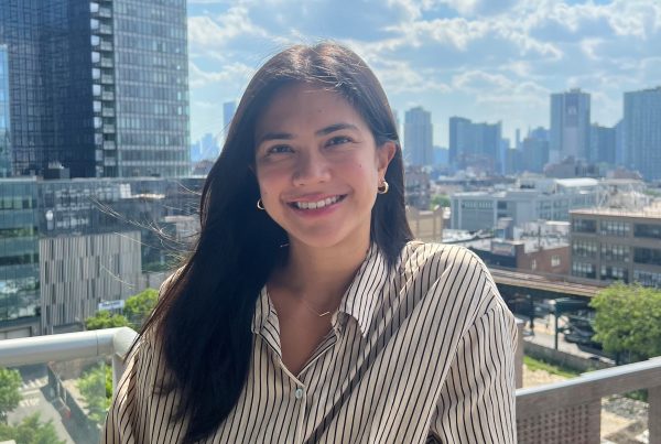 Women with dark hair standing on a balcony with a city view behind her, she is wearing a white and black striped shirt