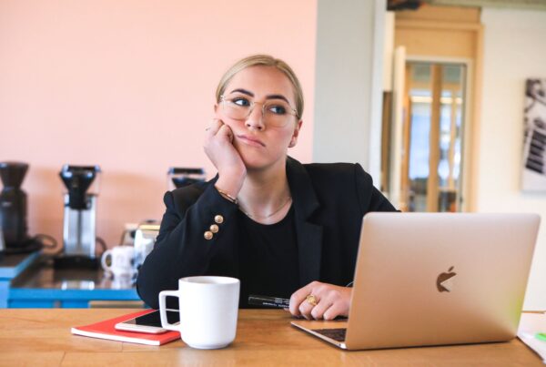 Young blonde women looking very bored sitting at a table with a laptop and cup in front of her